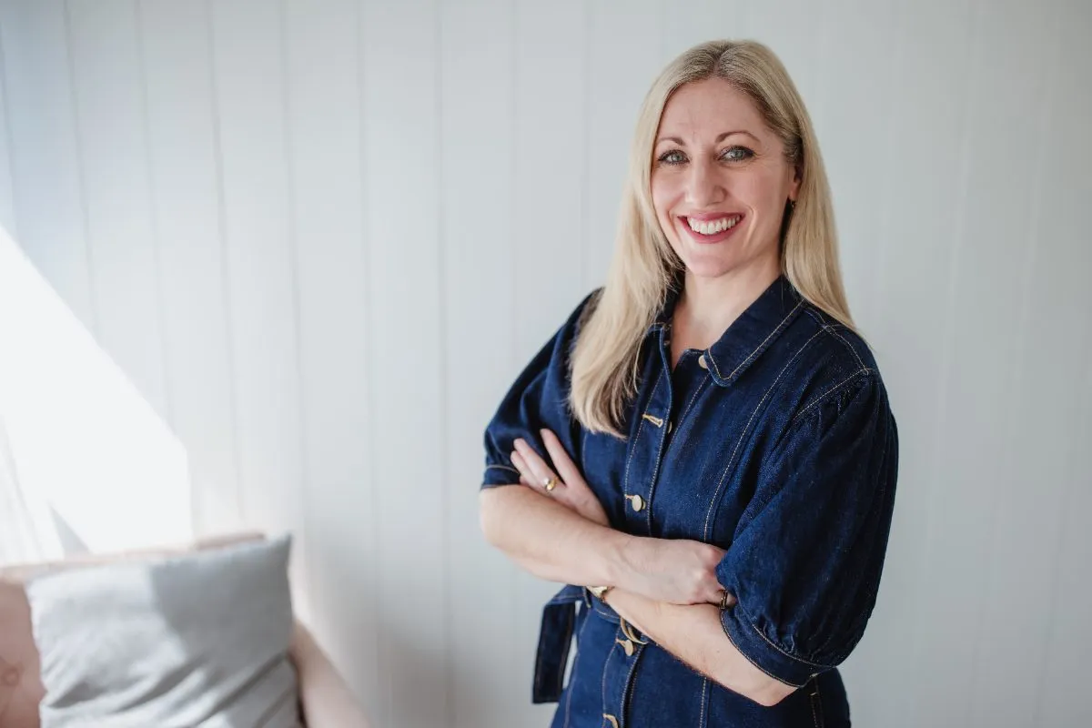 Kristy standing with arms crossed in a navy denim dress, smiling at the camera